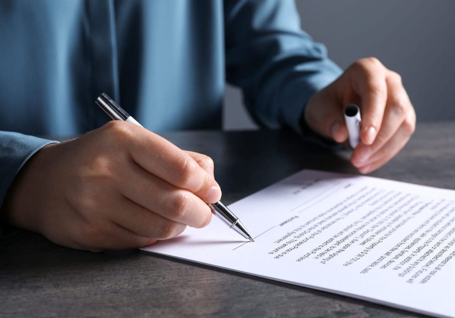 A person in a blue shirt signs a document with a pen on a dark table.